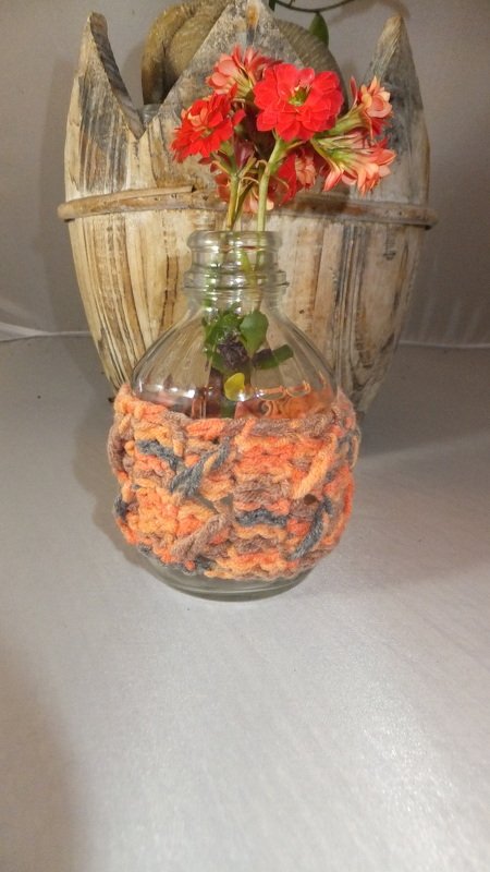 Glasvase mit gehäkelter orange-grauer Hülle und roten Blumen vor rustikaler Holzdeko