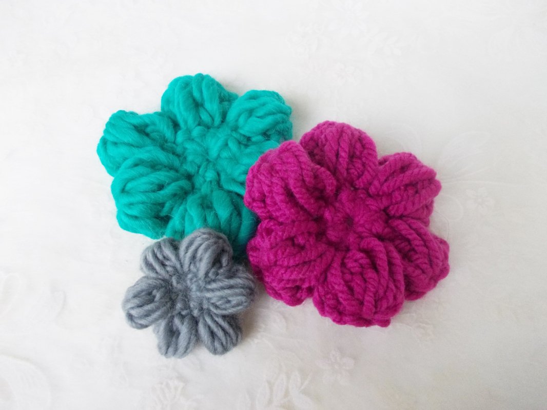 Drei gehäkelte Blumenapplikationen in Türkis, Magenta und Grau auf weißem Untergrund.