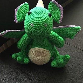 Gehäkelter grüner Amigurumi-Drache mit lila Akzenten und hellem Bauch, sitzend.
