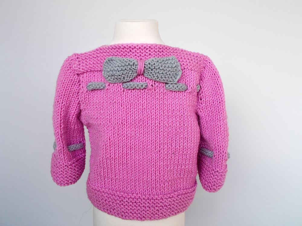 Rückenansicht eines rosa gestrickten Kinderpullovers mit großer grauer Schleife am Rücken und kleinen grauen Schleifen an den Ärmeln.