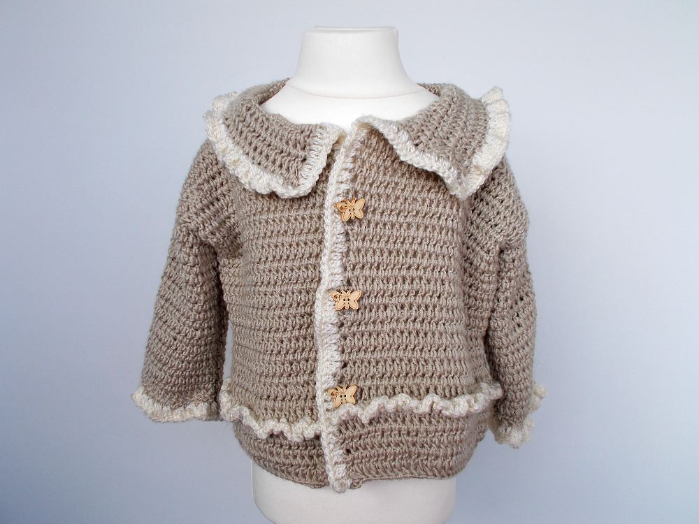 Beige gehäkelte Babyjacke mit weißer Rüsche und Schmetterlingsknöpfen auf Schaufensterpuppe