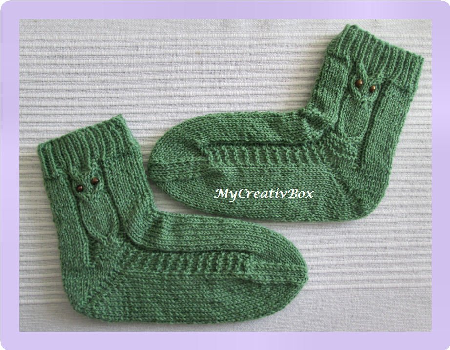 Paar grüne Babysocken mit Eulen-Zopfmuster und Knopfaugen auf hellem Hintergrund