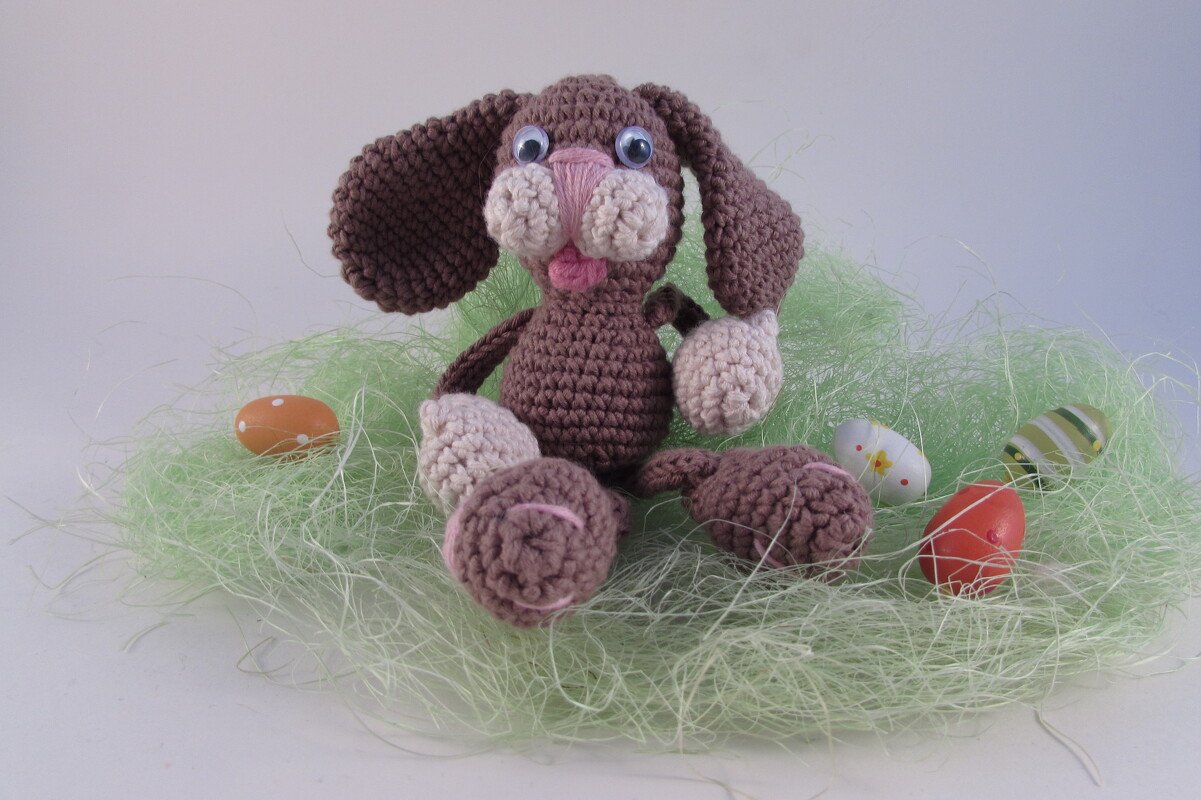 Gehäkelter brauner Amigurumi-Hase mit beigen Pfoten und Wackelaugen im grünen Ostergras neben bunten Ostereiern