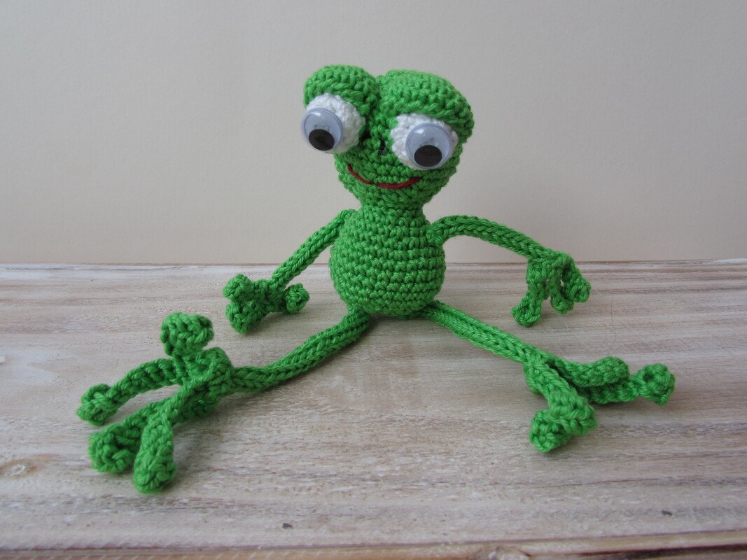 Grüner gehäkelter Amigurumi-Frosch mit großen Wackelaugen und rotem Lächeln