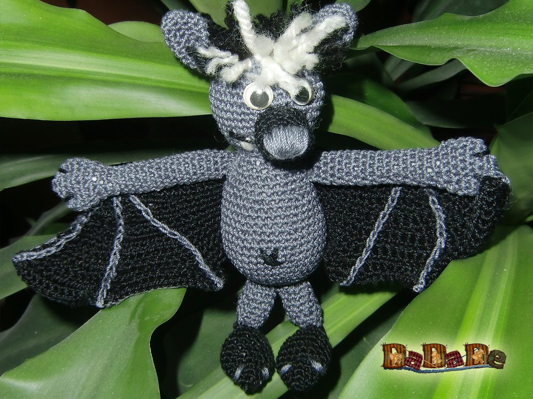 Grau-schwarze gehäkelte Fledermaus-Amigurumi mit weißen Haarbüscheln und Wackelaugen vor grünen Blättern