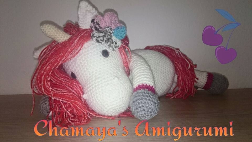 Weißes gehäkeltes Einhorn Amigurumi mit pinker Mähne und Blumenapplikation, liegend auf Holz.