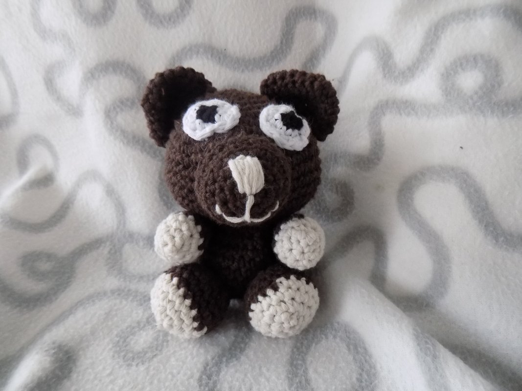 Kleiner dunkelbrauner Amigurumi-Teddybär mit weißen Akzenten an Augen, Schnauze, Pfoten und Füßen auf einer hellen, gemusterten Decke.