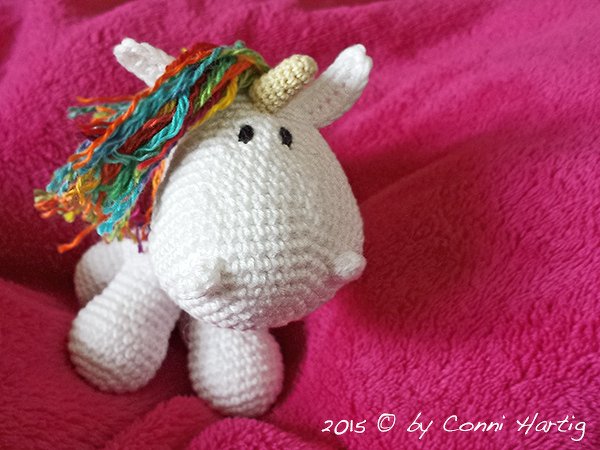 Weißes gehäkeltes Einhorn mit bunter Regenbogenmähne auf rosa Decke.
