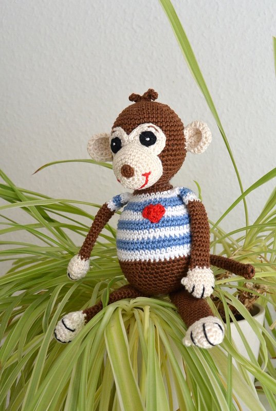 Gehäkelter brauner Amigurumi-Affe mit blau-weiß gestreiftem Shirt und rotem Herz auf grüner Pflanze
