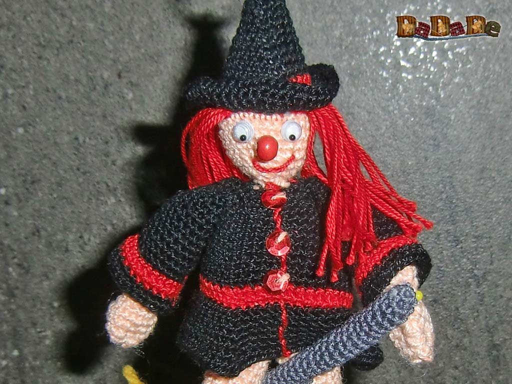 Handgehäkelte Hexenfigur mit rotem Haar, schwarzem Spitzhut, roter Nase und großen Augen, die einen kleinen Besen hält