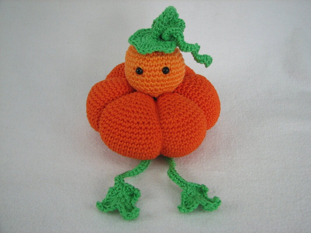 Gehäkelter orangefarbener Kürbis-Amigurumi mit schwarzen Augen und grünen Blättern auf weißem Hintergrund