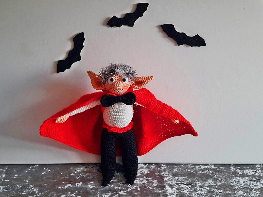 Gehäkelte Vampirpuppe mit rotem Umhang und schwarzer Fliege vor weißer Wand mit Fledermaus-Dekor