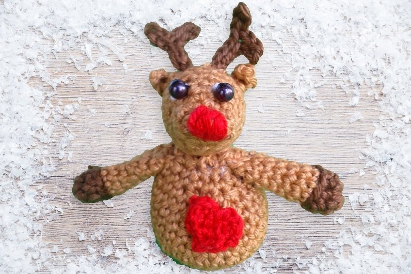 Gehäkeltes braunes Rentier mit roter Nase, lila Augen und rotem Herz auf schneebedecktem Holz