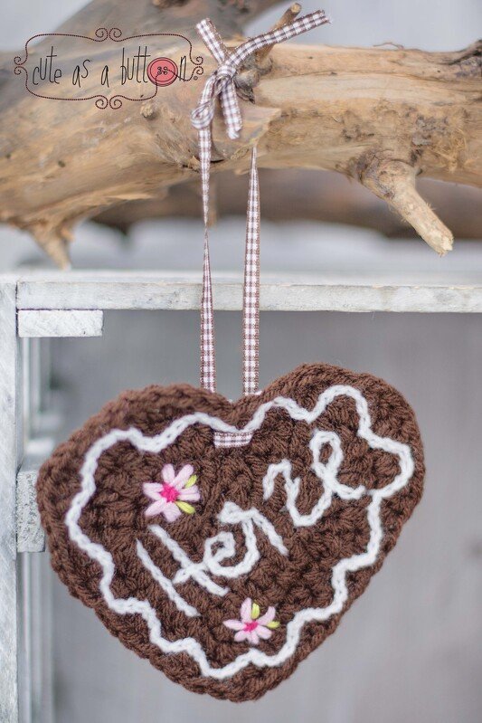 Braunes Häkelherz im Lebkuchen-Stil mit weißem 'Herz'-Schriftzug, rosa Blumen und kariertem Band