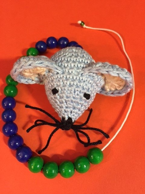 Hellblauer gehäkelter Amigurumi‑Mauskopf mit bestickten Augen und Perlenring auf rotem Hintergrund