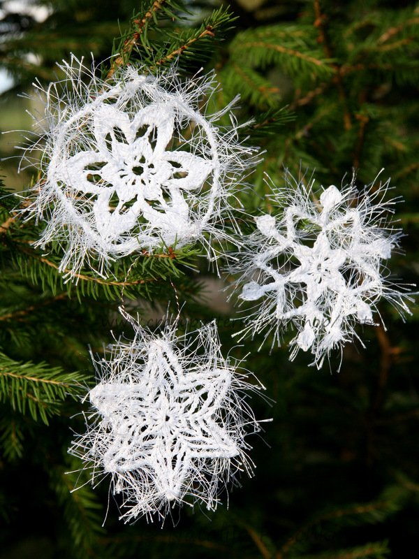 Drei weiße, flauschige gehäkelte Schneeflocken und Sterne an grünen Tannenzweigen