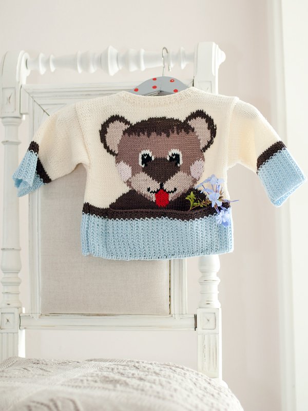Gestrickter Baby-Pullover creme und hellblau mit eingestricktem Bärenmotiv an weißem Stuhl