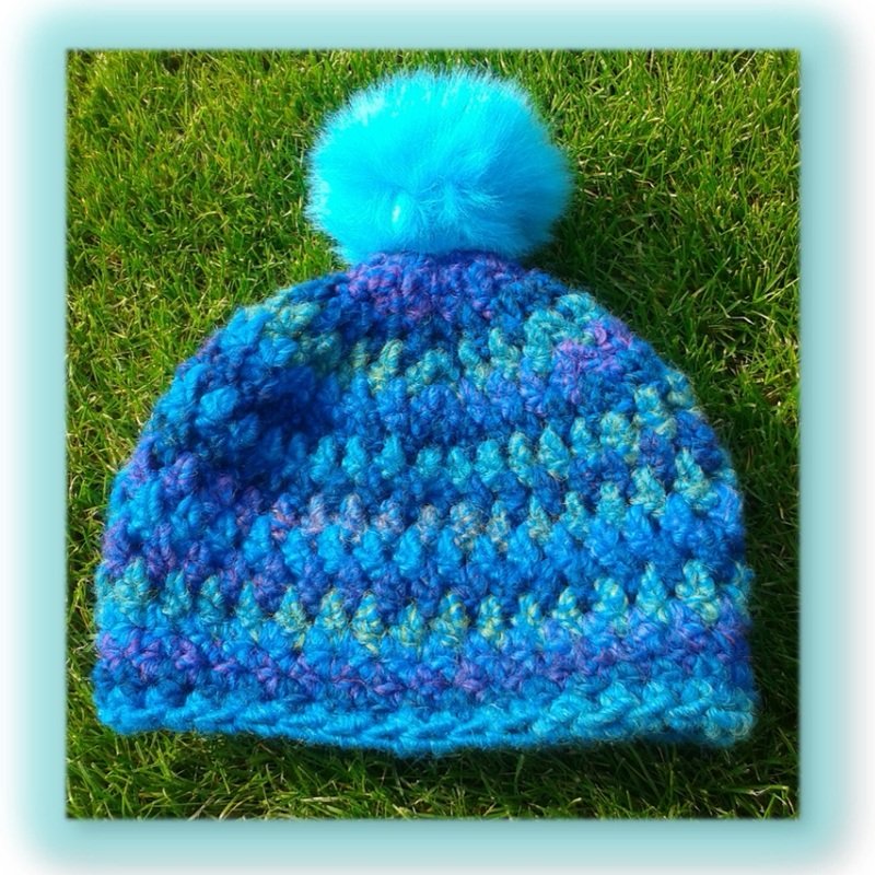 Gehäkelte blau-türkise Mütze mit flauschigem Bommel auf Rasen