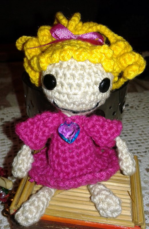 Gehäkelte Amigurumi Puppe mit gelben Locken, schwarzen Augen und pinkem Kleid, sitzt auf Holzunterlage
