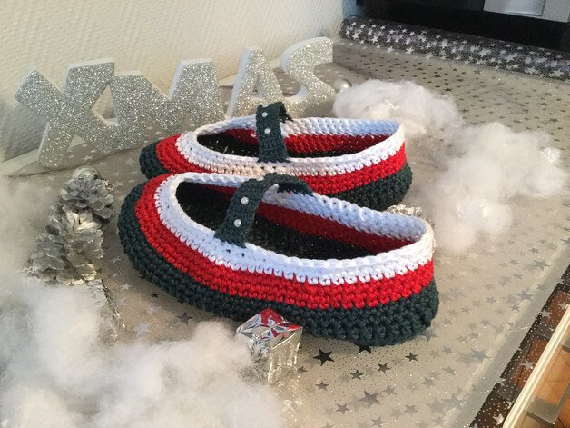 Ein Paar gehäkelter Hausschuhe in Rot, Dunkelgrün und Weiß mit Riemchen und Perlen auf weihnachtlich dekorierter Fläche mit Kunstschnee.