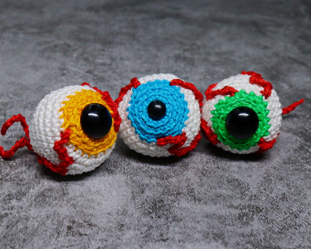 Drei gehäkelte Augen mit gelber, blauer und grüner Iris, schwarzen Pupillen und roten Garnadern auf dunklem Untergrund.