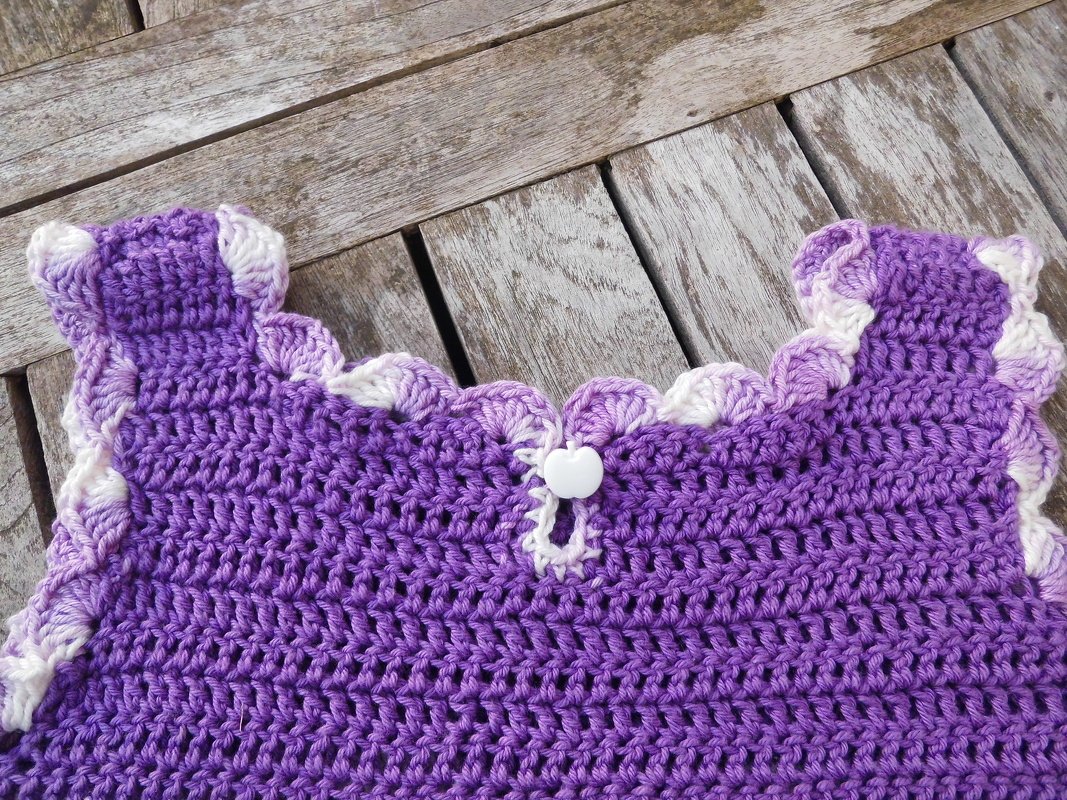 Gehäkeltes lila Oberteil mit weißem Muschelrand und kleinem weißem Knopf auf Holztisch