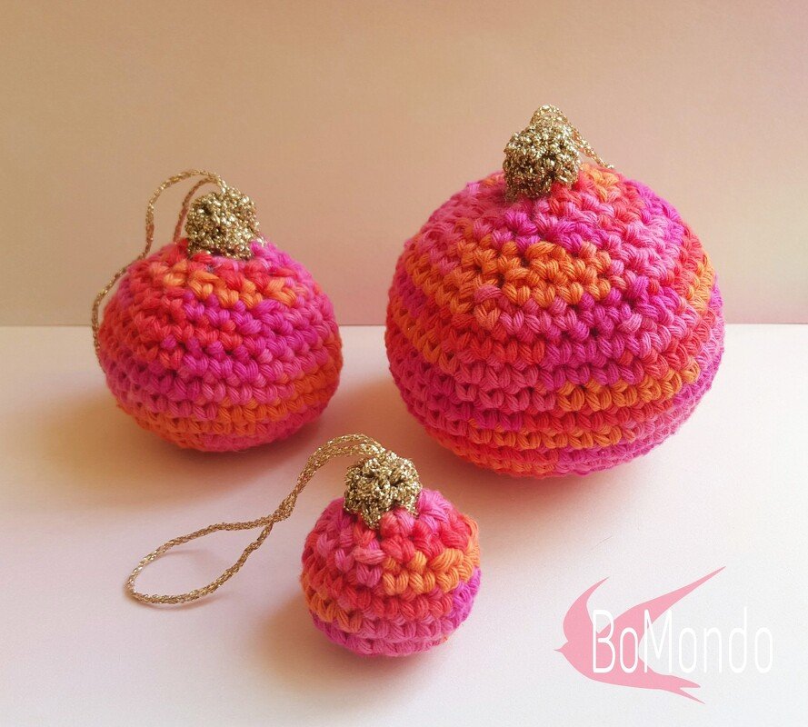Drei gehäkelte Kugeln in Pink und Orange mit goldenen Aufhängschlaufen auf hellem Untergrund