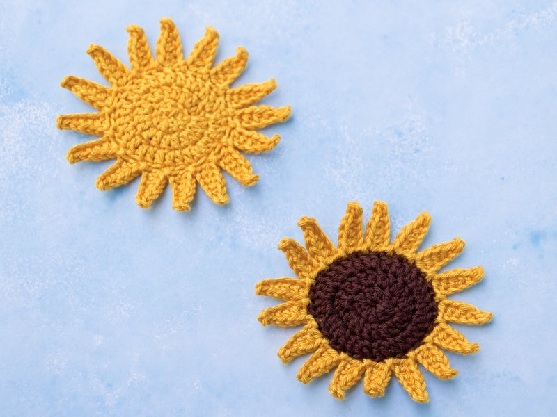Zwei runde, gehäkelte Applikationen auf blauem Hintergrund: Eine gelbe Sonne und eine gelb-braune Sonnenblume.