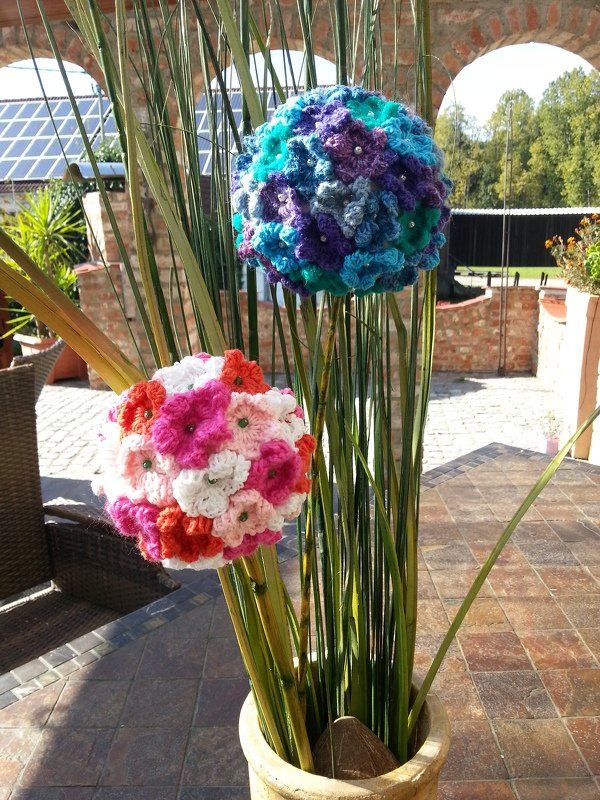 Zwei runde, gehäkelte Blumenkugeln in einem hellen Pflanzkübel auf einer Terrasse; oben blau‑türkis‑violett, unten rosa‑weiß‑orange; lange grüne Stiele.