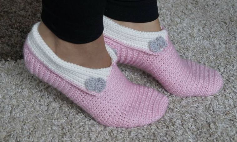 Gehäkelte rosa Haussocken mit weißer Umschlagmanschette und grauer Blütenapplikation an den Füßen einer Person