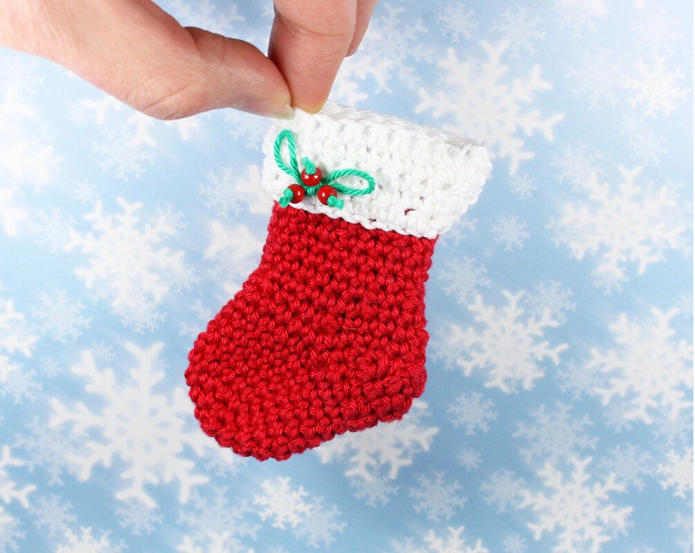 Hand holding a small red and white crocheted Christmas stocking with green yarn loops and red bead holly detail on a pale blue snowflake background.