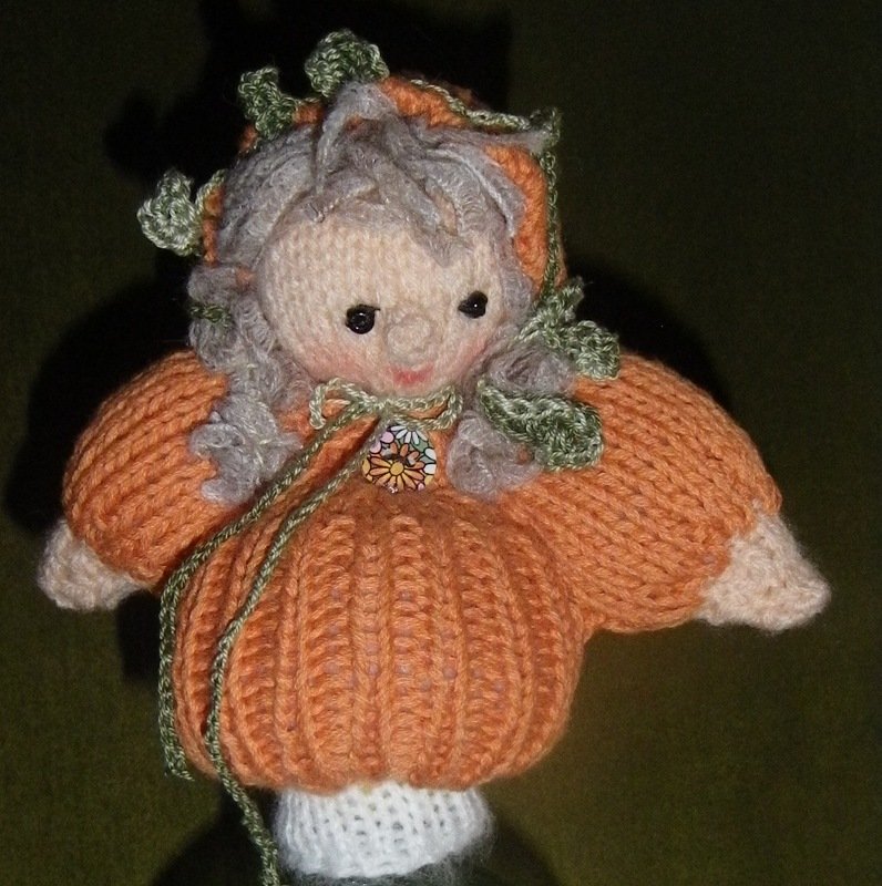 Gestrickte Kinderpuppe im orangefarbenen Kürbiskleid mit grauen Locken und grünen Blättern