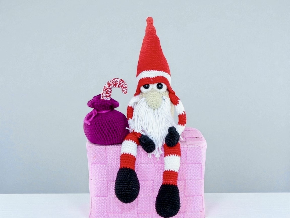 Crochet Christmas gnome with tall red hat, white beard and striped legs sitting on a pink woven box beside a purple sack