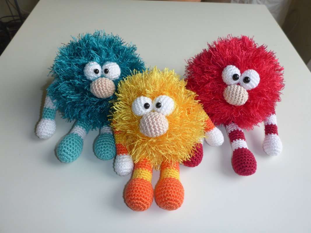 Drei gehäkelte Amigurumi-Monster in Türkis, Gelb und Rot mit großen Augen und gestreiften Gliedmaßen.