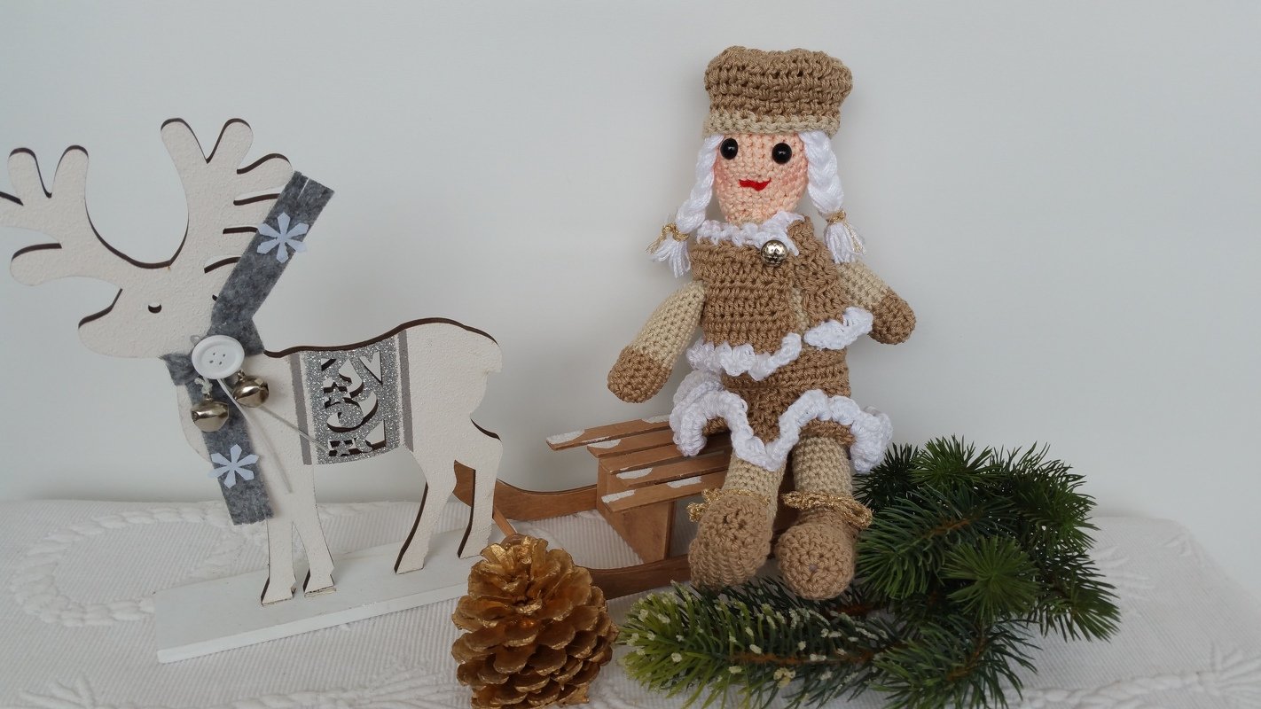 Gehäkelte Amigurumi-Puppe in Braun und Weiß auf Holzschlitten neben Holzrentier, Tannenzapfen und Tannenzweigen.