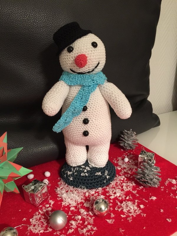 Gehäkelter Schneemann mit schwarzem Zylinder, blauem Schal und roter Nase auf rotem Filztuch mit Kunstschnee