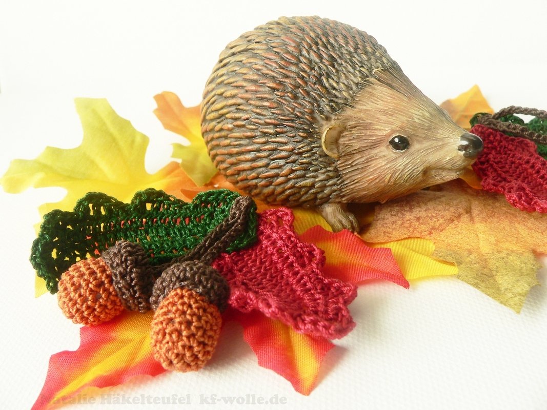 Igel-Dekofigur mit gehäkelten roten und grünen Blättern sowie gehäkelten Eicheln auf herbstlichem Laub