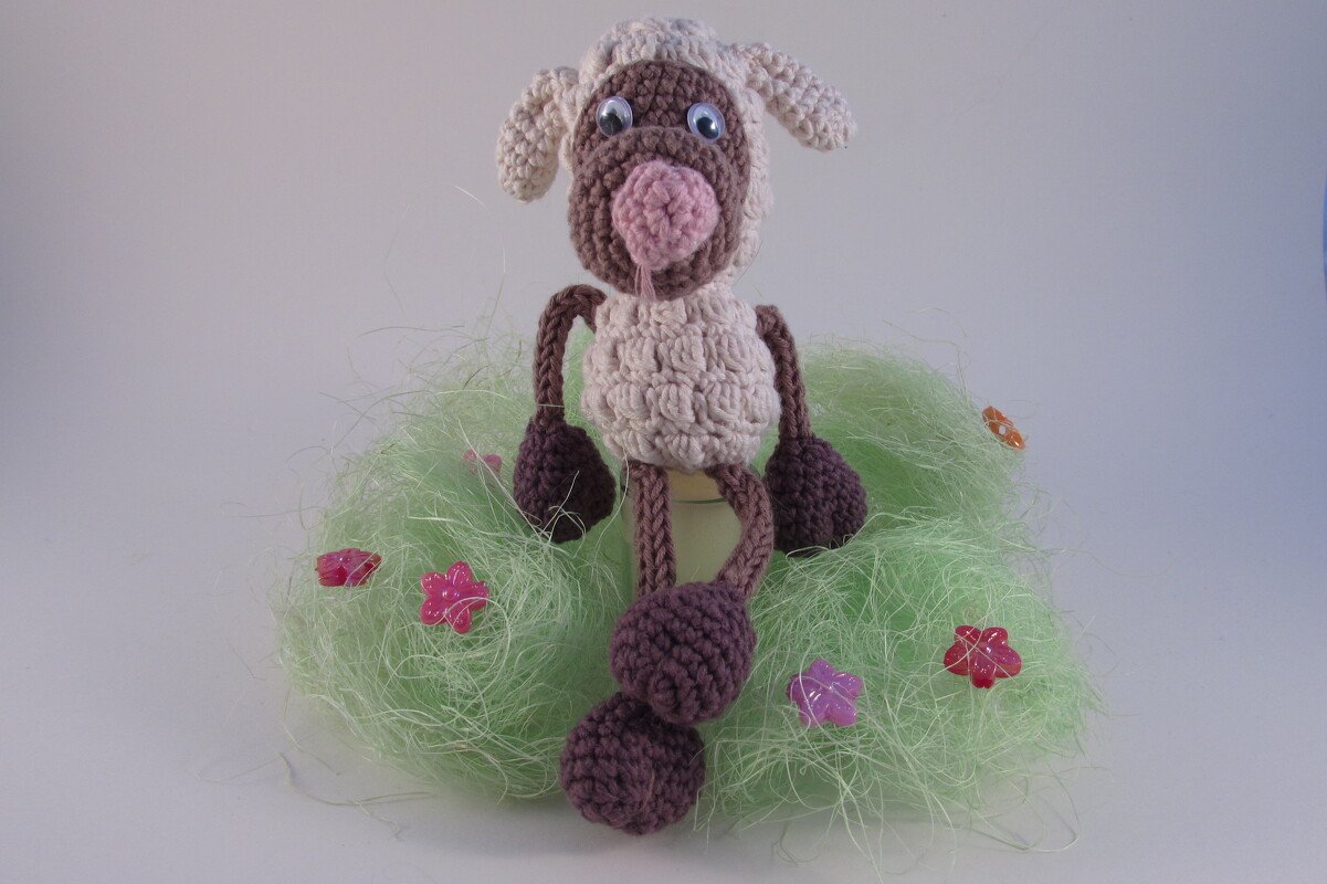 Gehäkeltes Amigurumi-Schaf mit strukturierter Körpermasche, gedrehten Kordelbeinen und Bommelenden auf grüner Deko-Faser