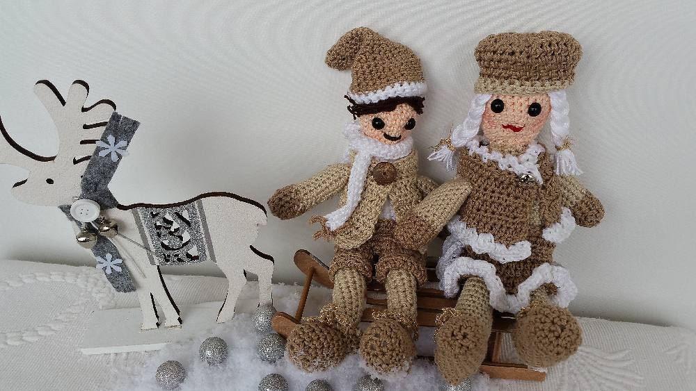 Zwei gehäkelte Amigurumi-Puppen in Winterkleidung auf einem Holzschlitten neben einem weißen Holzrentier
