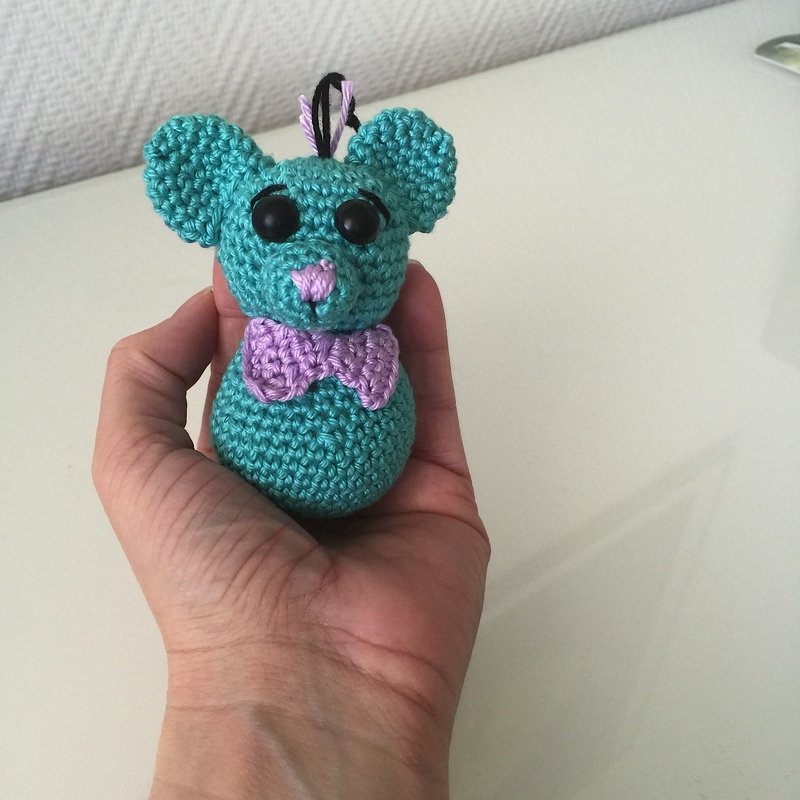 Gehäkelte türkise Amigurumi-Maus mit lila Schleife, gehalten in einer Hand