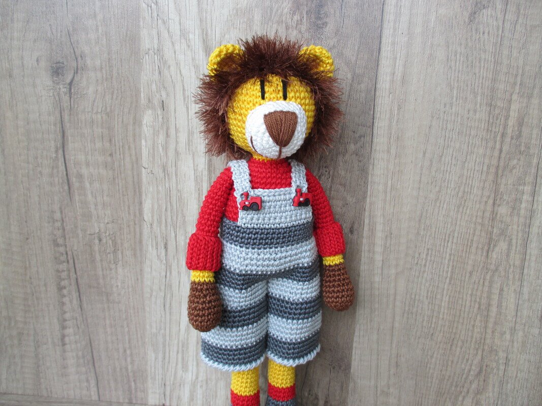 Gehäkelte Löwenpuppe mit brauner Mähne, rotem Shirt und grau-dunkelgrau gestreifter Latzhose vor Holzuntergrund