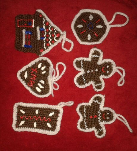 Sechs gehäkelte Lebkuchen-Anhänger in Braun mit weißem Rand und farbigen Stickdetails auf rotem Hintergrund