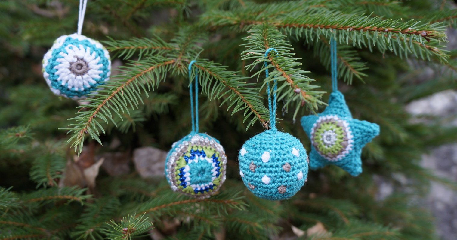 Vier handgehäkelte Weihnachtsornamente in Blau-, Grün- und Weißtönen an einem Tannenzweig.