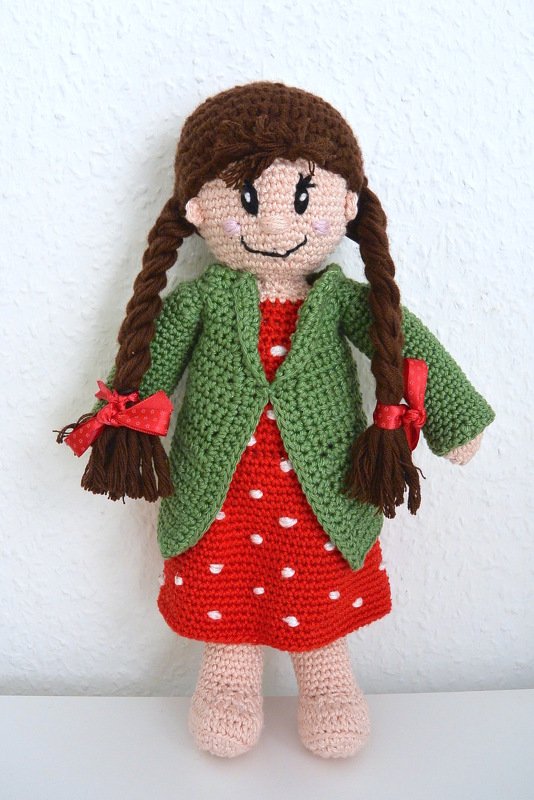 Gehäkelte Amigurumi-Puppe mit braunen Zöpfen, rotem gepunktetem Kleid und grüner Jacke.