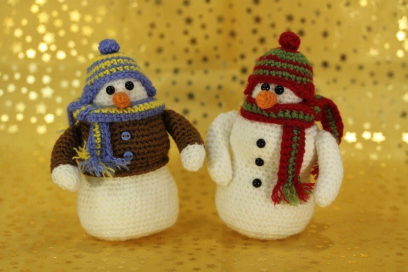 Zwei gehäkelte Amigurumi-Schneemänner mit Mützen und Schals vor goldenem Sternhintergrund, einer mit braunem Pullover.