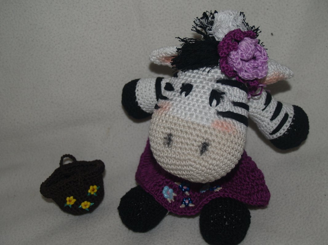 Gehäkeltes Zebra-Amigurumi in Weiß und Schwarz mit lila Kleid, Blüte am Kopf und brauner Tasche mit gelben Blumen.