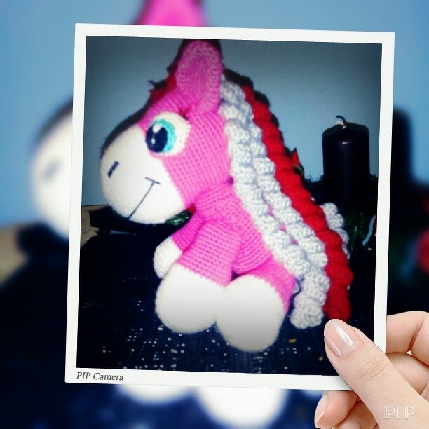 Rosa gehäkeltes Einhorn‑Amigurumi mit blauem Auge und rot‑weißer Lockenmähne.