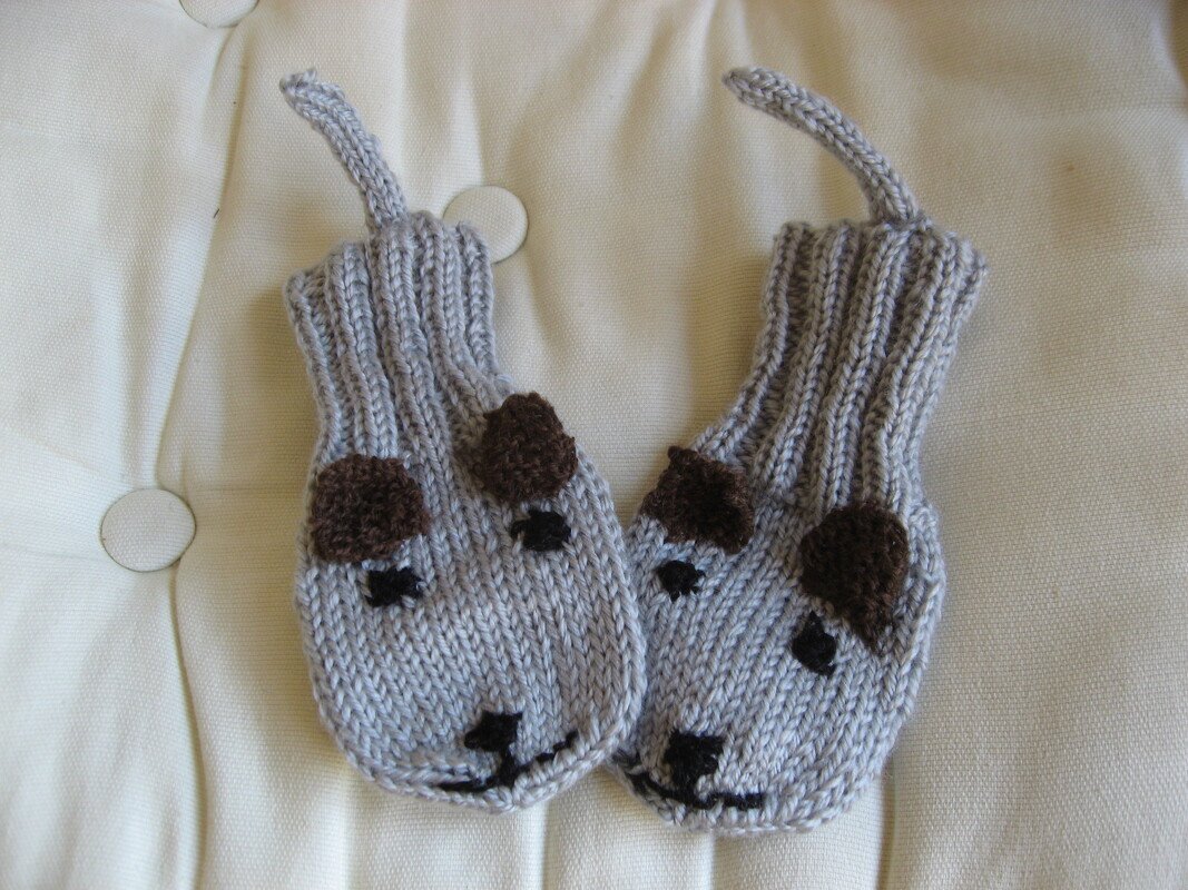 Zwei hellgraue, gestrickte Baby-Fäustlinge mit gestickten Hundegesichtern und braunen Ohren auf gepolsterter hellbeiger Oberfläche.