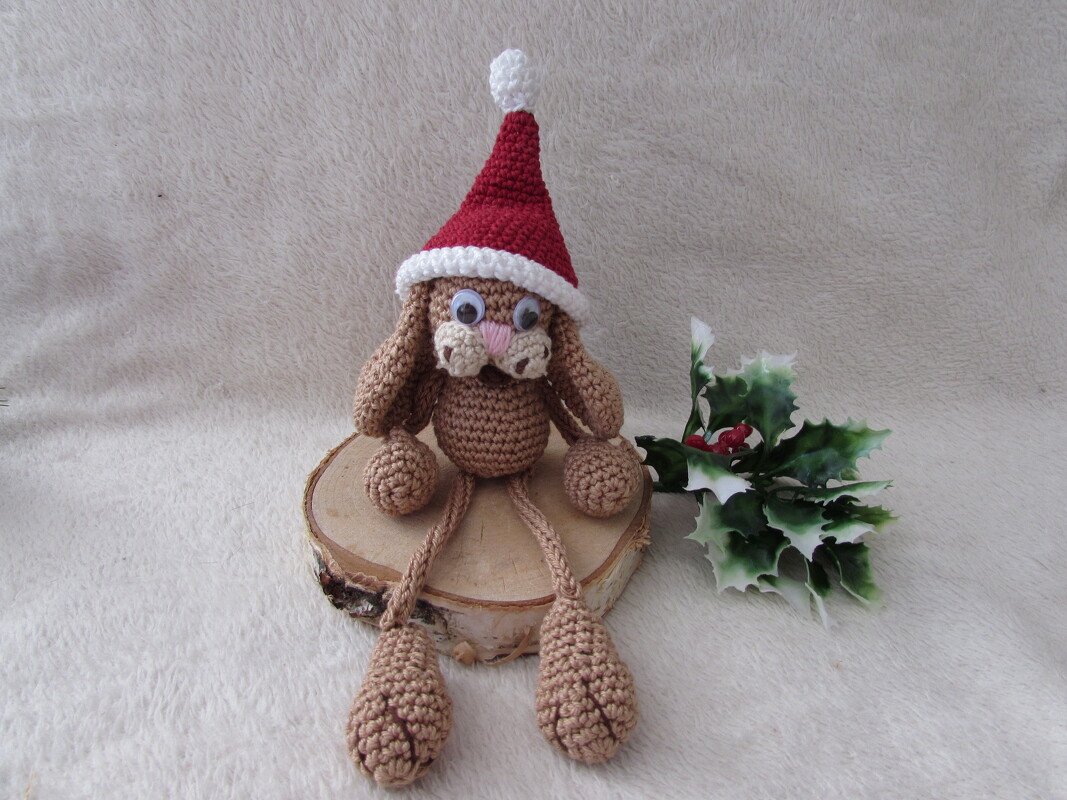Amigurumi‑Hase mit langen Beinen und roter Nikolausmütze auf Holzscheibe