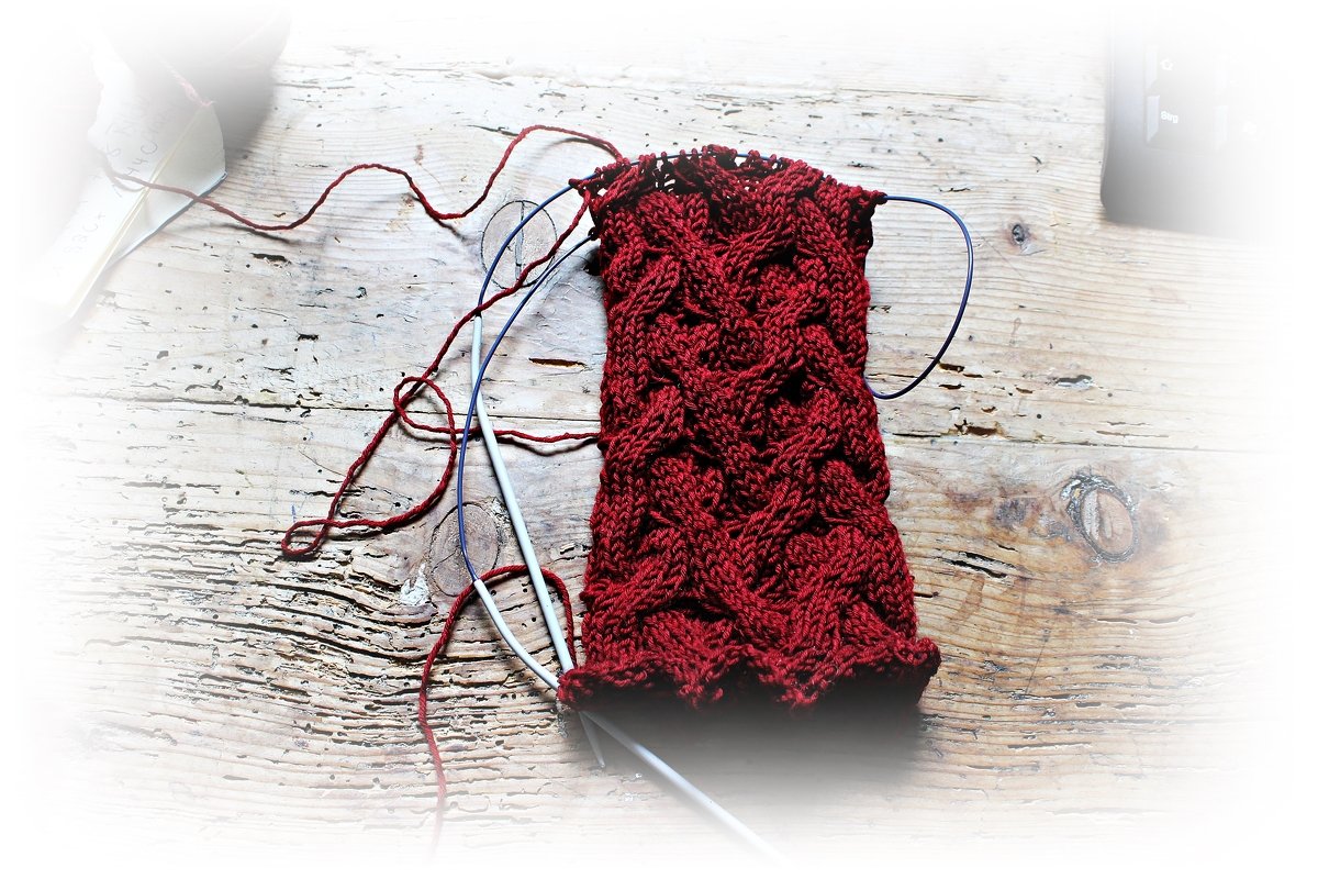 Nahaufnahme dunkelroter Armstulpe mit Zopfmuster und Rundstricknadeln auf Holztisch